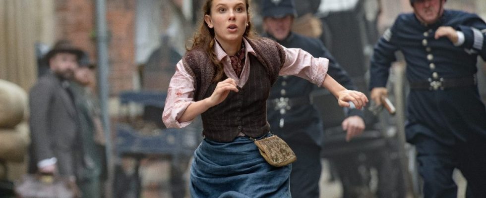 A press image of Millie Bobby Brown as Enola being chased by cops down a street in Enola Holmes 2.