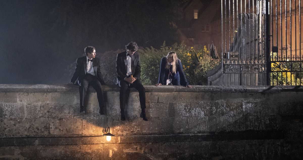 Oliver (Barry Keoghan) et Felix Catton (Jacob Elordi), en smoking, sont assis ensemble sur un petit pont de pierre au-dessus d'un étang avec Venetia (Alison Oliver) à proximité à Saltburn.