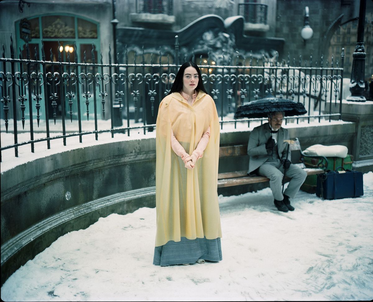 Emma Stone dans le rôle de Bella Baxter se tient dans la neige, avec un Mark Ruffalo déprimé sous un parapluie derrière elle dans Poor Thigns