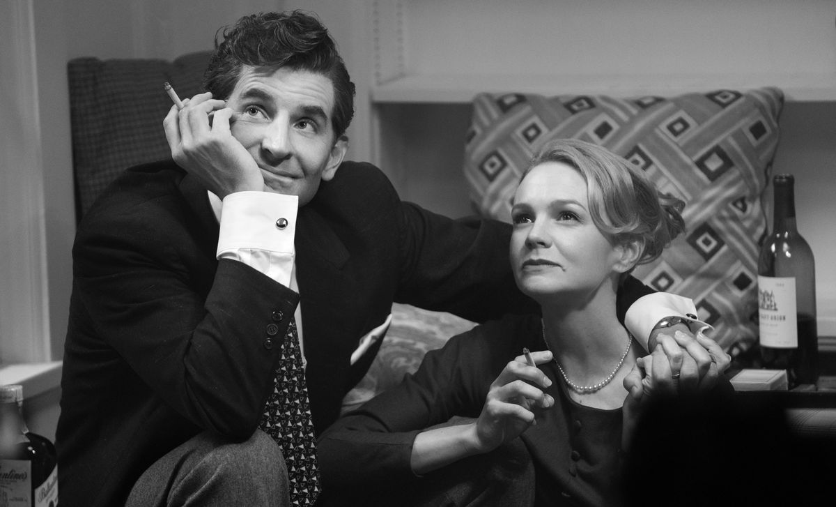 Bradley Cooper dans le rôle de Leonard Bernstein et Carey Mulligan dans le rôle de Felicia Montealegre sont assis ensemble, fumant et souriant à quelqu'un hors écran, dans une image en noir et blanc de Maestro de Netflix.
