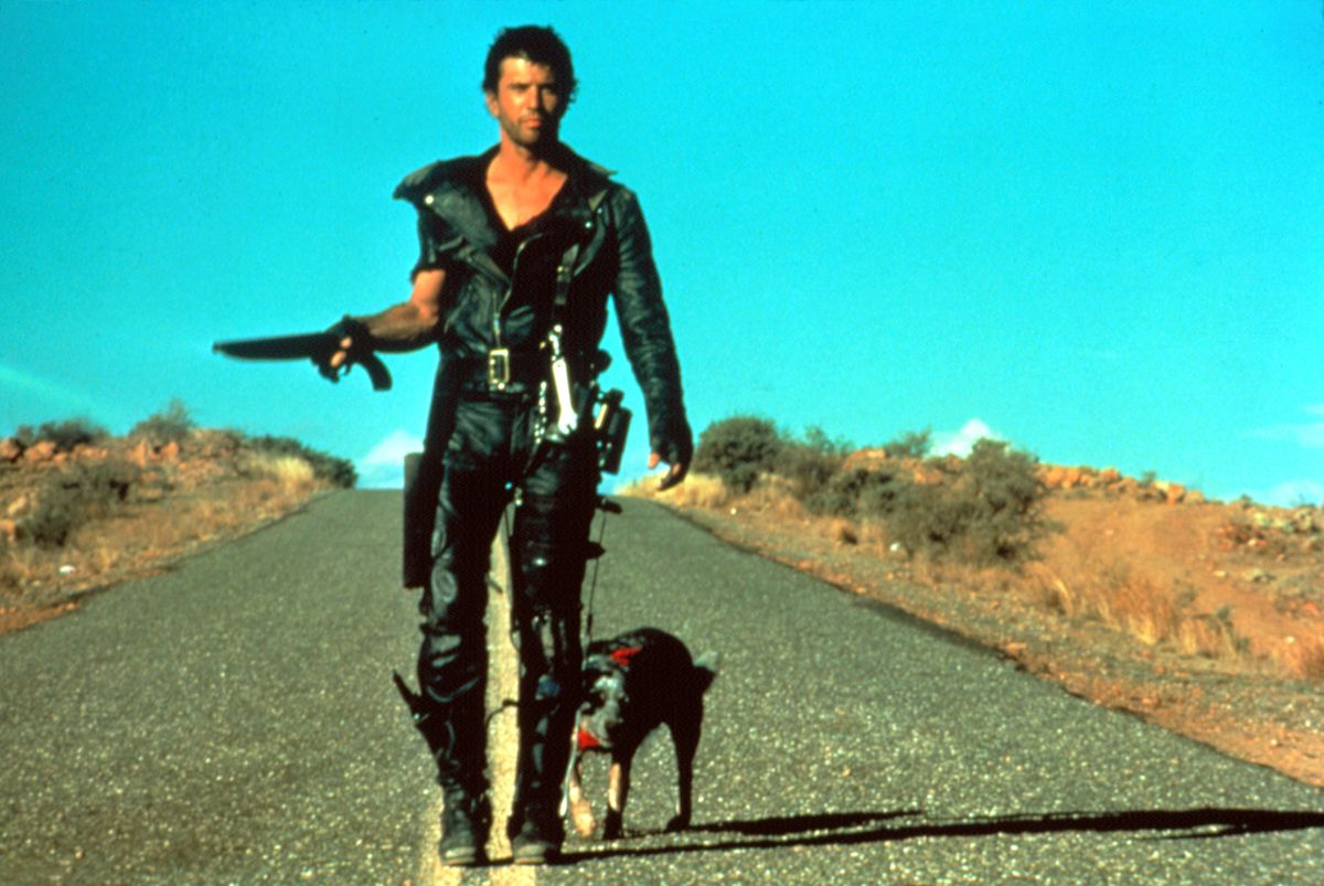 Mel Gibson dans le rôle de Mad Max, vêtu de cuir noir et tenant un petit fusil de chasse, se promène avec un chien sur une route dans Mad Max 2 : The Road Warrior.