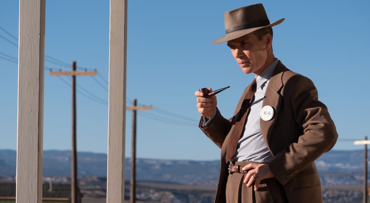 J. Robert Oppenheimer (Cillian Murphy), en costume marron et chapeau, tient une pipe et se tient dans un désert près d'une rangée de poteaux téléphoniques dans Oppenheimer de Christopher Nolan