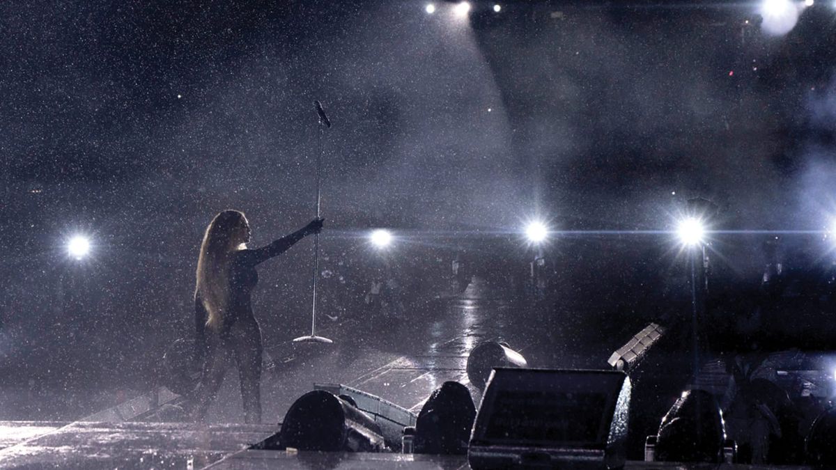 Beyoncé tient un micro debout sur scène pendant la tournée mondiale Renaissance, alors que des lignes lumineuses brillent et que la pluie tombe.