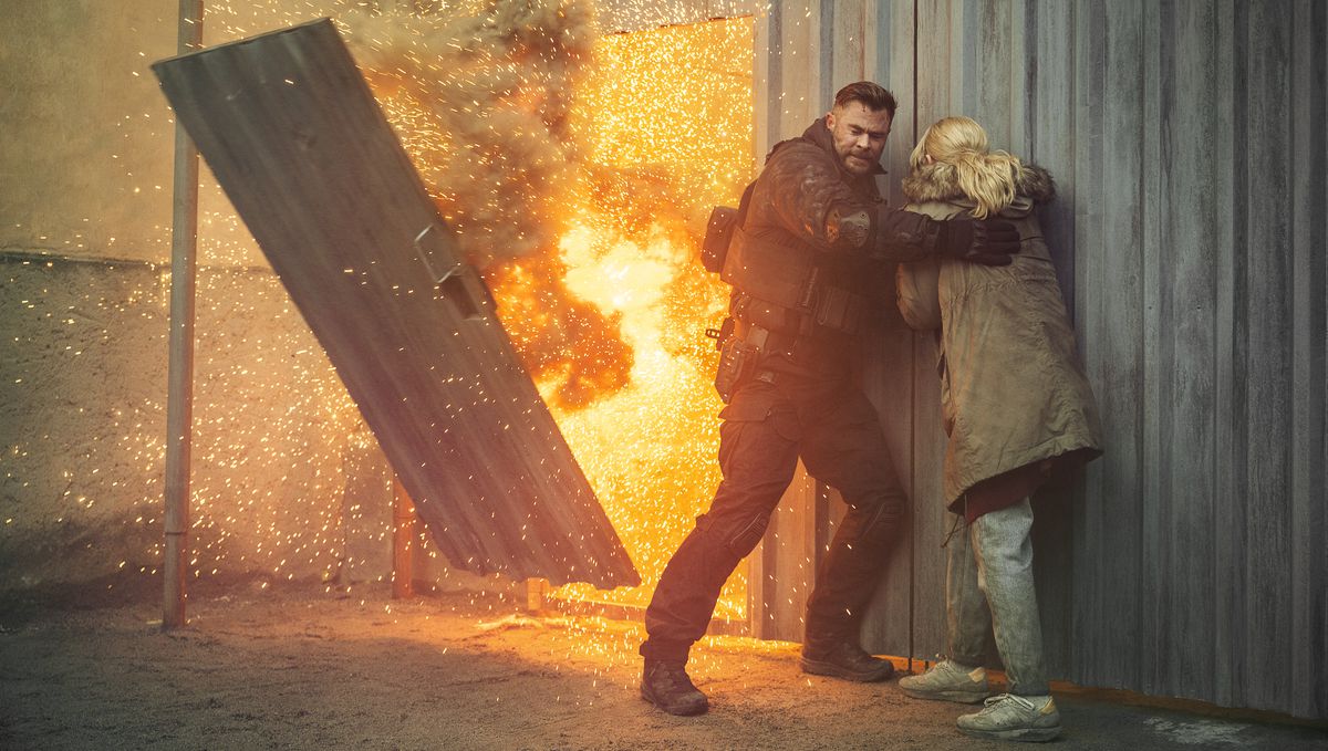Le mercenaire Tyler Rake (Chris Hemsworth) et sa belle-sœur Ketevan (Tinatin Dalakishvili) s'abritent contre un mur de tôle ondulée alors qu'une porte de ce mur s'ouvre de manière explosive dans Extraction 2.