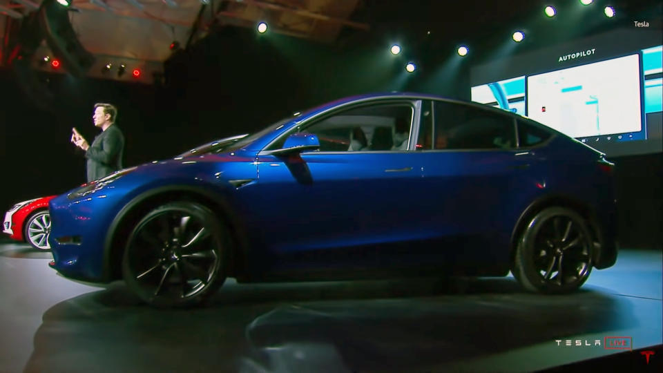 Un véhicule électrique Tesla Model Y bleu est assis sur scène lors d’un événement de 2019.  Elon Musk est vu derrière, s'adressant à un public depuis une vue de profil par rapport à la caméra.