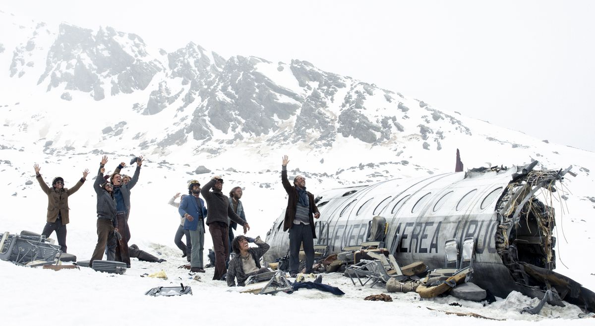 Un groupe de survivants, vus comme de minuscules personnages à une longue distance, se tiennent dans la neige à côté de leur avion fragmenté et saluent quelque chose hors écran dans la Société de la Neige de Netflix.
