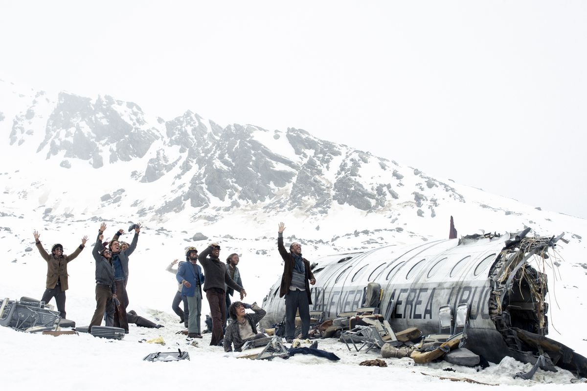 Un groupe d’hommes et de femmes debout à côté de l’épave d’un avion avec une montagne enneigée en arrière-plan dans La Société de la Neige.