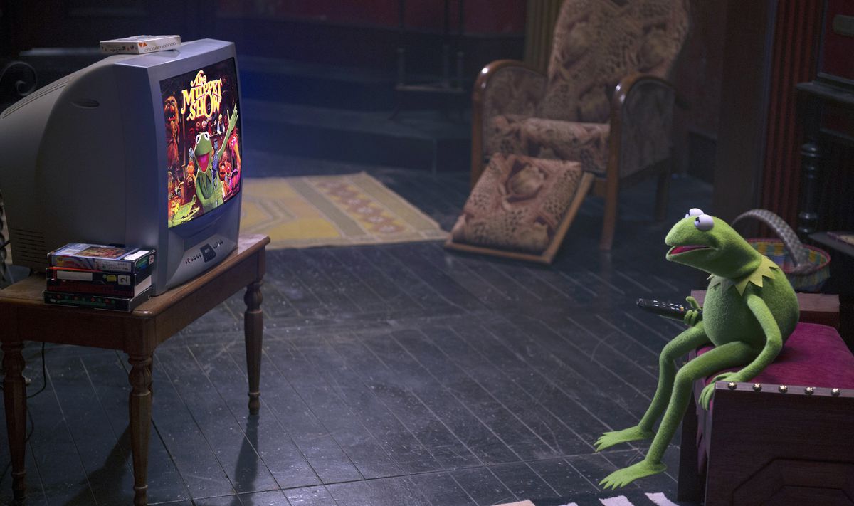 Kermit la grenouille est assise sur un banc bas rembourré rouge et regarde le Muppet Show à la télévision dans Muppets Most Wanted