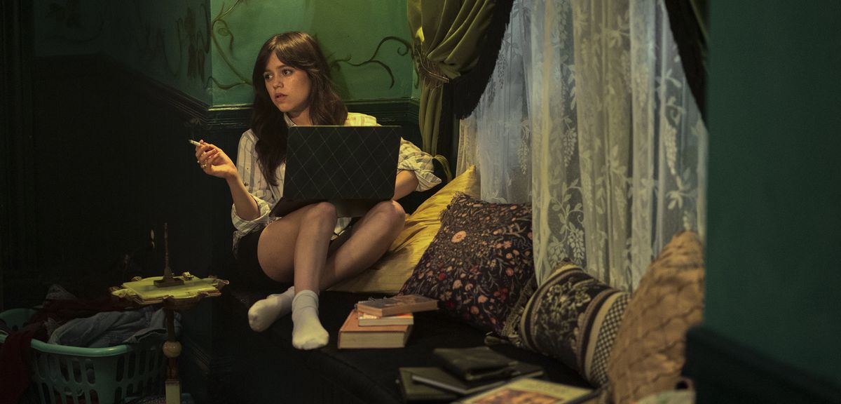 Le Caire (Jenna Ortega) est assise et fume avec un ordinateur portable sur ses genoux et ses jambes repliées sur une chaise, entourée d'oreillers, de livres et d'un rideau de dentelle vaporeux dans Miller's Girl