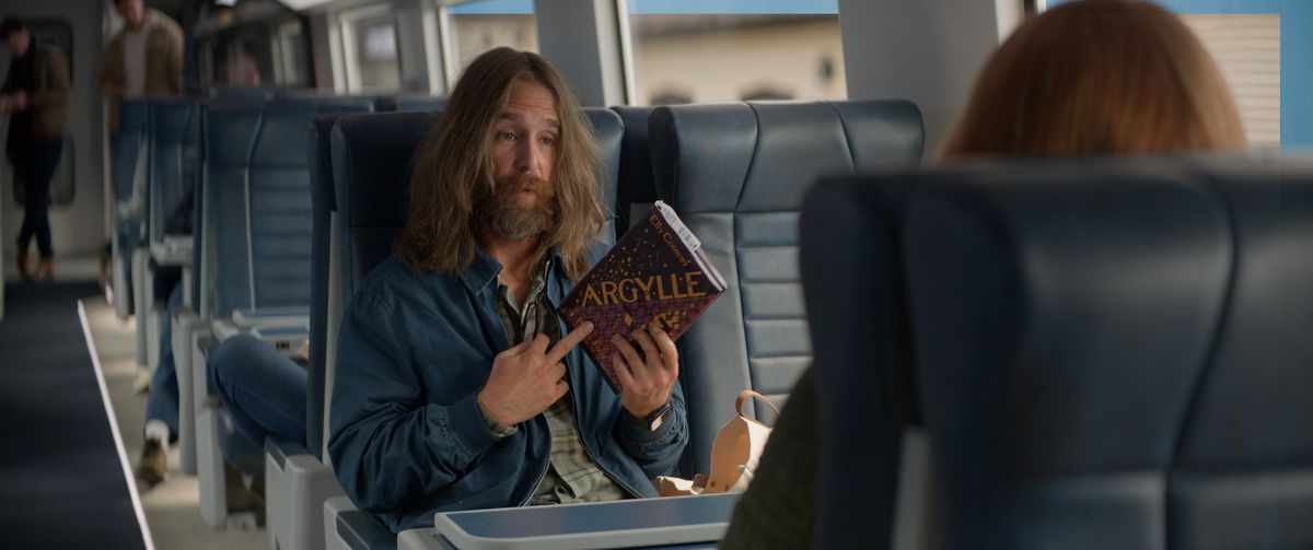 Sam Rockwell, aux cheveux longs et à la barbe, tient un exemplaire du livre Argylle d'Elly Conway dans le film Argylle