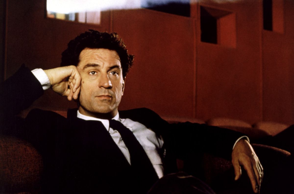 Le réalisateur hollywoodien David Merrill (un jeune Robert De Niro, en costume-cravate noir et chemise à col blanc) est assis dans une salle de cinéma aux murs rouges et regarde un film avec la lumière du projecteur derrière lui dans Guilty by Suspicion de 1991.