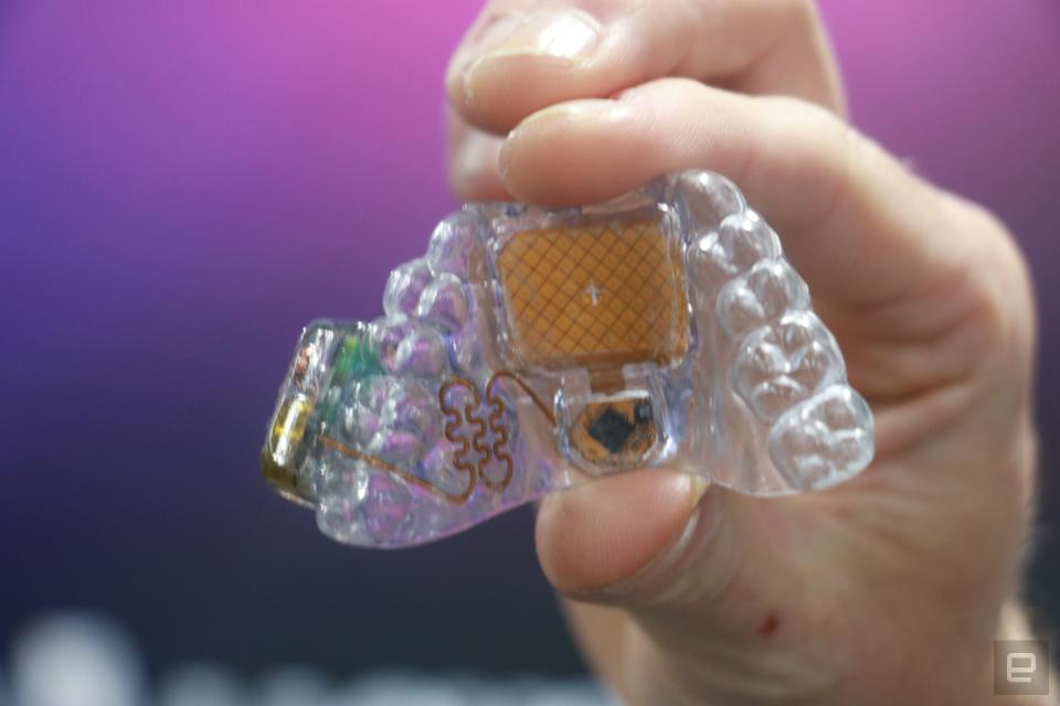 Le MouthPad, un contrôleur à commande vocale, tenu dans les airs.  Il s'agit d'un plateau dentaire transparent avec un pavé tactile orange au milieu et des circuits partout.