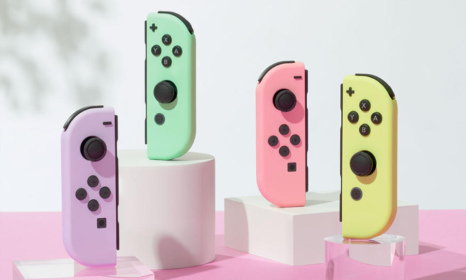 Photo marketing de deux paires de manettes Nintendo Switch Joy-Con aux couleurs pastel.  De gauche à droite : violet (contrôleur gauche), vert (contrôleur droit), rose (contrôleur gauche) et jaune (contrôleur droit).  Ils sont assis debout sur des supports sur une table rose sur fond blanc.