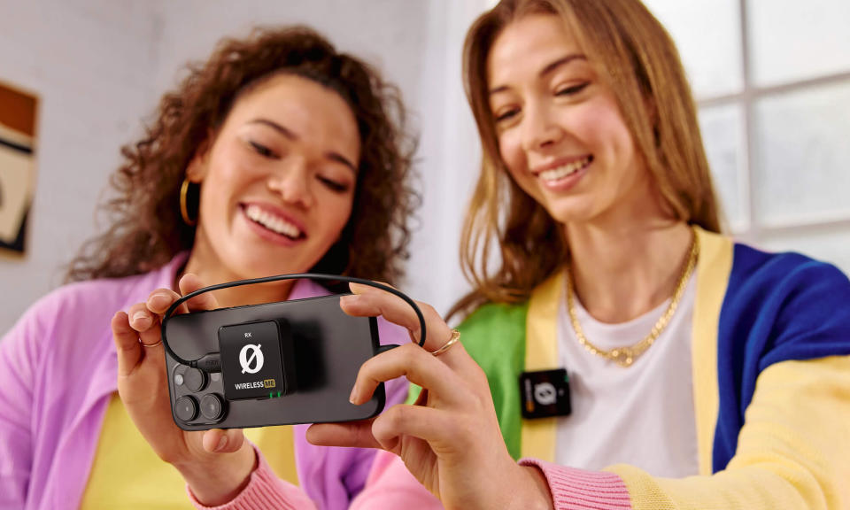 Photo marketing de style de vie du produit pour le micro Rode Wireless ME.  Deux jeunes sourient en regardant un téléphone doté d'un récepteur fixé à l'arrière.  L’un porte un micro sans fil.