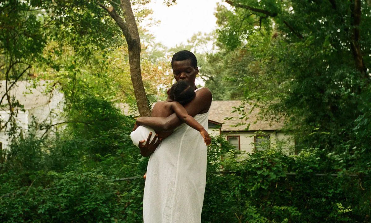Une femme (Sheila Atim) en robe blanche berçant un enfant dans ses bras devant un bosquet d'arbres et de buissons avec une maison en arrière-plan dans All Dirt Roads Taste of Salt.