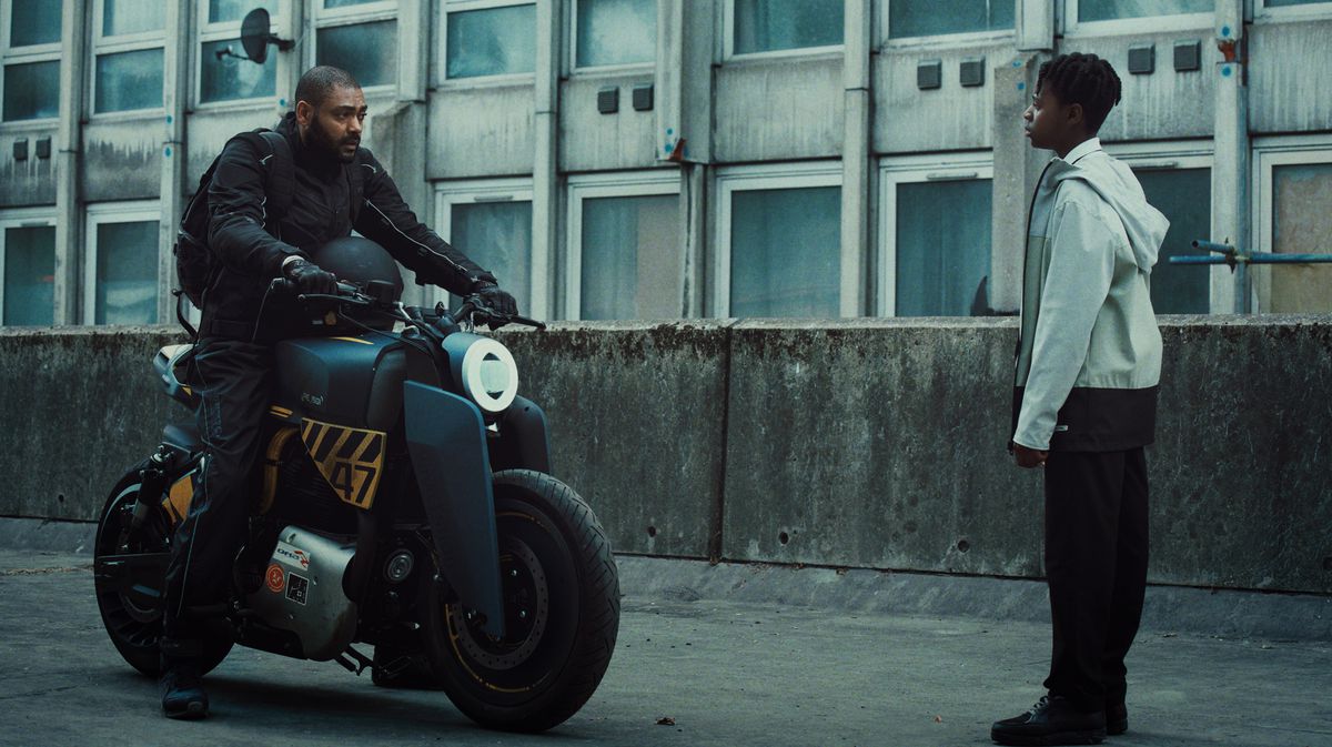 (LR) Un homme en survêtement noir (Kane Robinson) au sommet d'une moto futuriste se tient devant un jeune garçon vêtu d'un sweat à capuche blanc (Jedaiah Bannerman) et d'un pantalon noir dans The Kitchen.