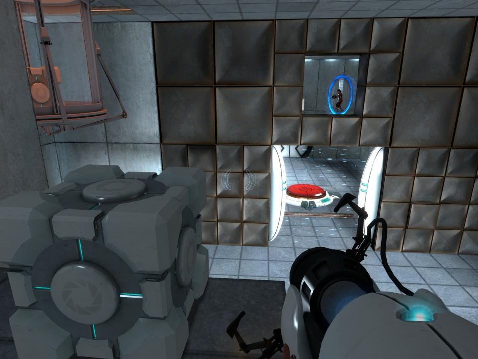 Gameplay toujours du jeu de réflexion Portal de Valve.  La perspective à la première personne du haut d'une plate-forme montre le joueur tenant le pistolet à portail alors qu'un cube se trouve à proximité.  Un interrupteur rouge se trouve au sol en arrière-plan derrière une porte.