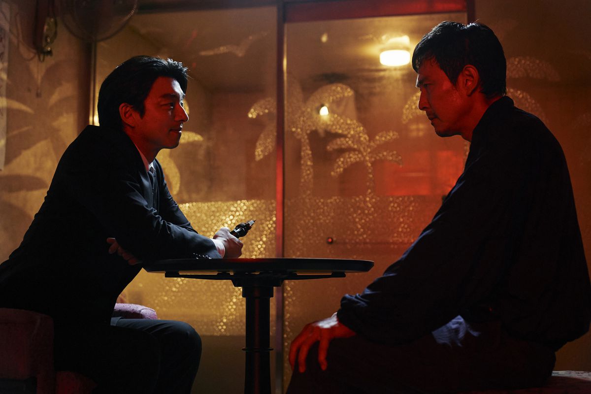 Le recruteur et Gi-hun (joués respectivement par Gong Yoo et Lee Jung-jae) assis à une table, le recruteur tenant ce qui ressemble à une arme pointée loin de la table.