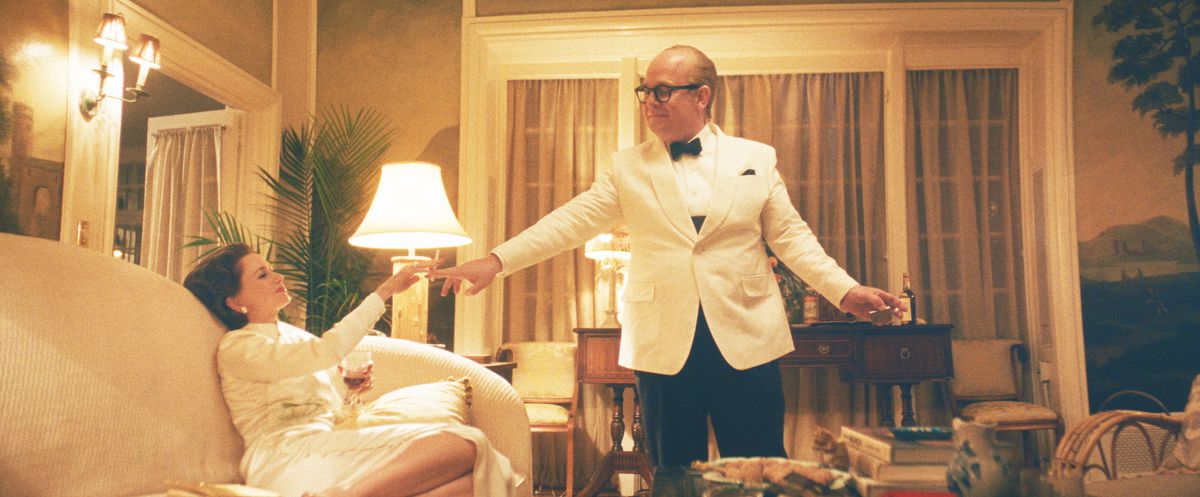 Capote, vêtu d'un smoking blanc et d'un nœud papillon, se tient au-dessus de Babe Paley, allongée sur un canapé vêtue d'une robe blanche, alors qu'il lui tend une cigarette dans Feud de FX : Capote contre les cygnes.