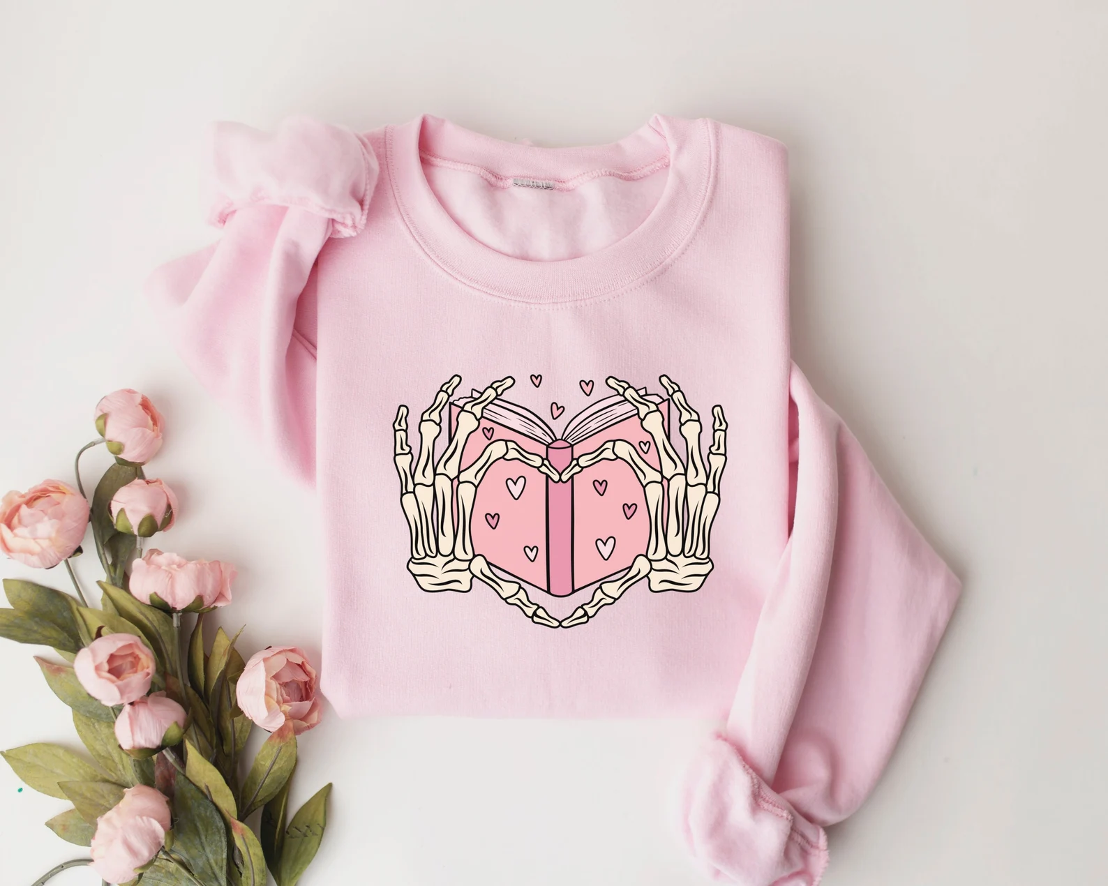 Image d'un sweat-shirt rose en forme de cœur réalisé par deux mains squelettes.  Entre les mains se trouve un livre. 
