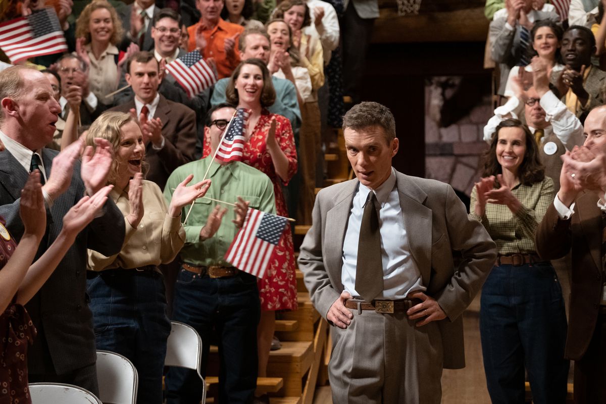 J. Robert Oppenheimer se tient debout, les mains sur la taille, alors qu'il marche entre des tribunes jumelles de gradins bondés remplis de gens applaudissant agitant des drapeaux américains à la main dans une scène du film Oppenheimer.