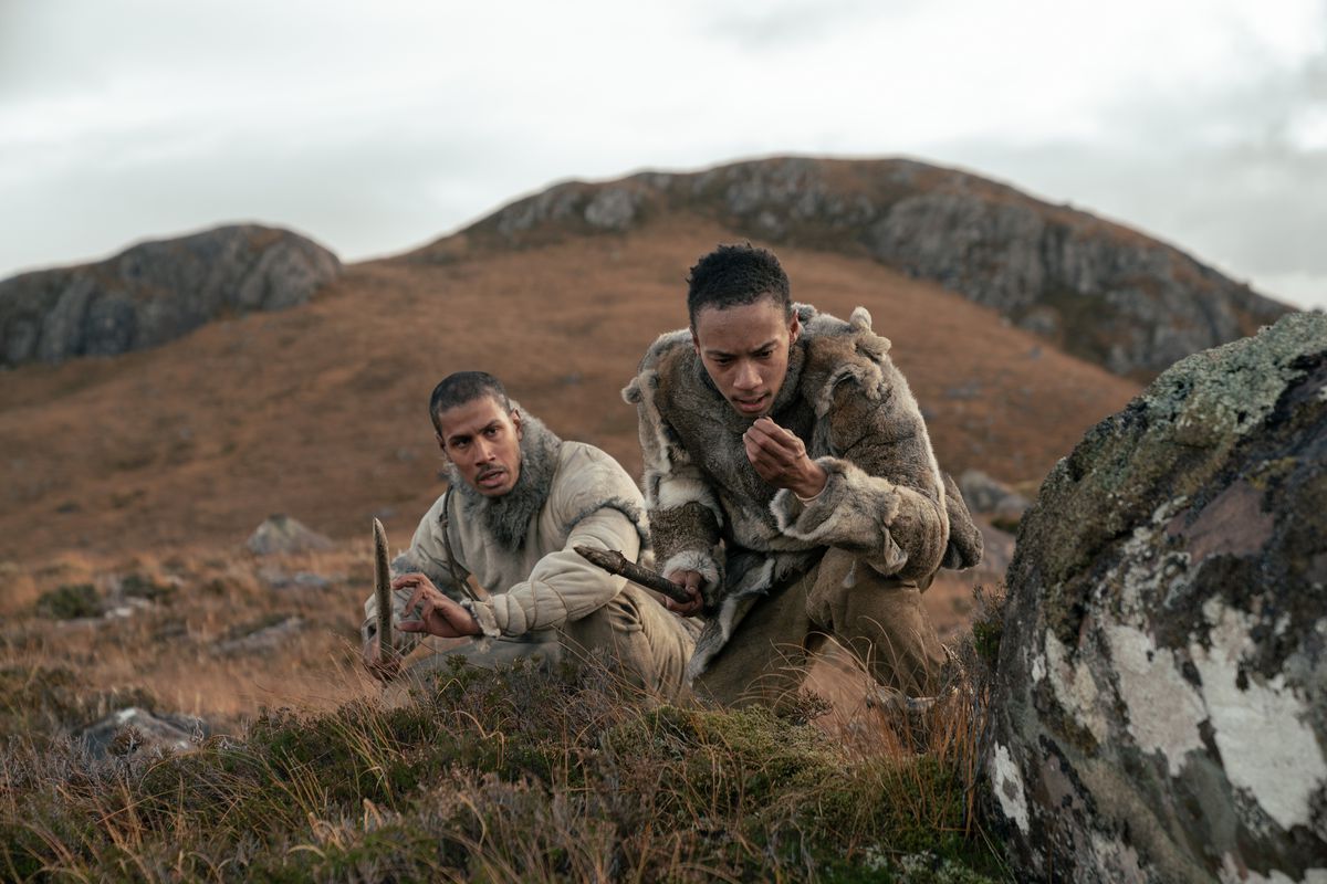 Deux hommes paléolithiques, Adem et Geirr (Chuku Modu et Kit Young), tiennent leurs lances alors qu'ils traquent quelque chose sur une colline brune, herbeuse et pierreuse dans Out of Darkness.