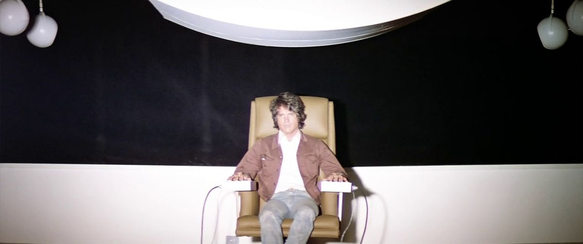 Warren Beatty dans le rôle de Lee Carter, assis sur une chaise blanche, regardant un écran lumineux.