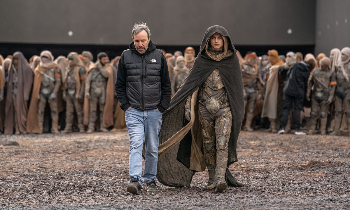 Denis Villeneuve et Timothée Chalamet en costume traversent un champ de gravier sur le tournage de Dune Part 2, un grand groupe de figurants costumés en Fremen derrière eux.