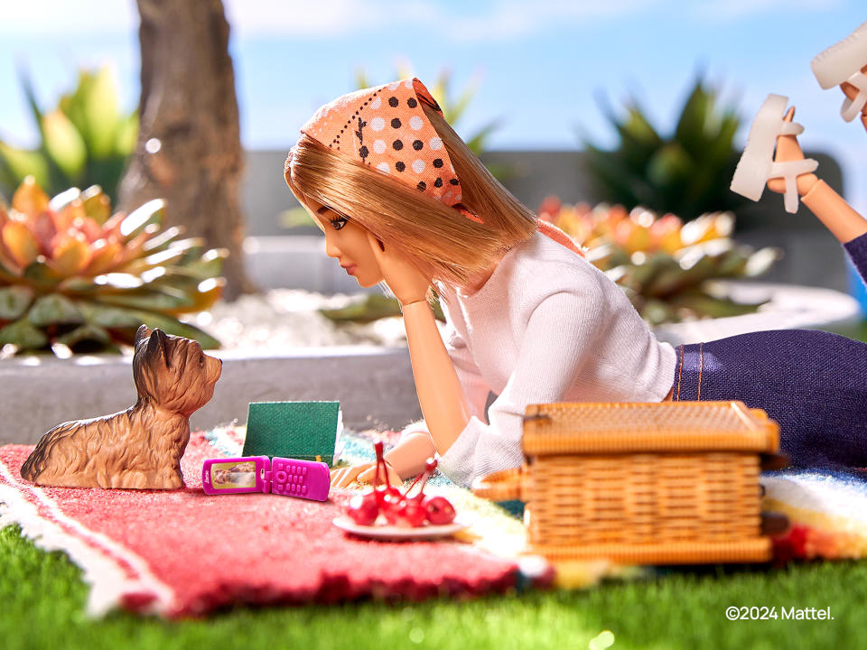 Une Barbie est représentée allongée sur une table de pique-nique avec un chien en plastique devant elle et un téléphone à clapet rose ouvert sur le côté.