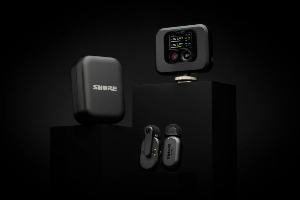 Photo du produit du pack Shure MoveMic Two.  Deux micros-cravates sans fil, un étui de chargement et un récepteur sont posés sur des socles noirs devant un fond noir spectaculaire.