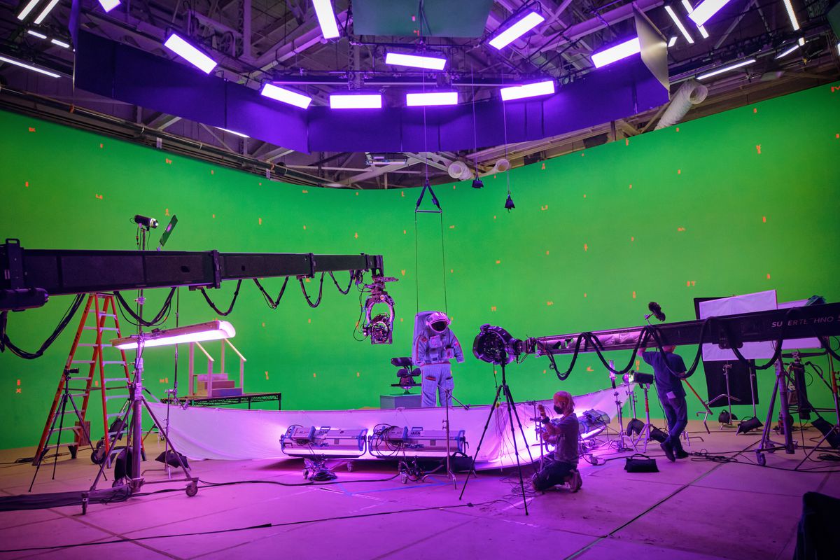 Une silhouette en combinaison spatiale est éclipsée par des grues, des caméras et des équipements dans un espace vert et violet aux hauts plafonds, sur le tournage de Spaceman de Netflix.