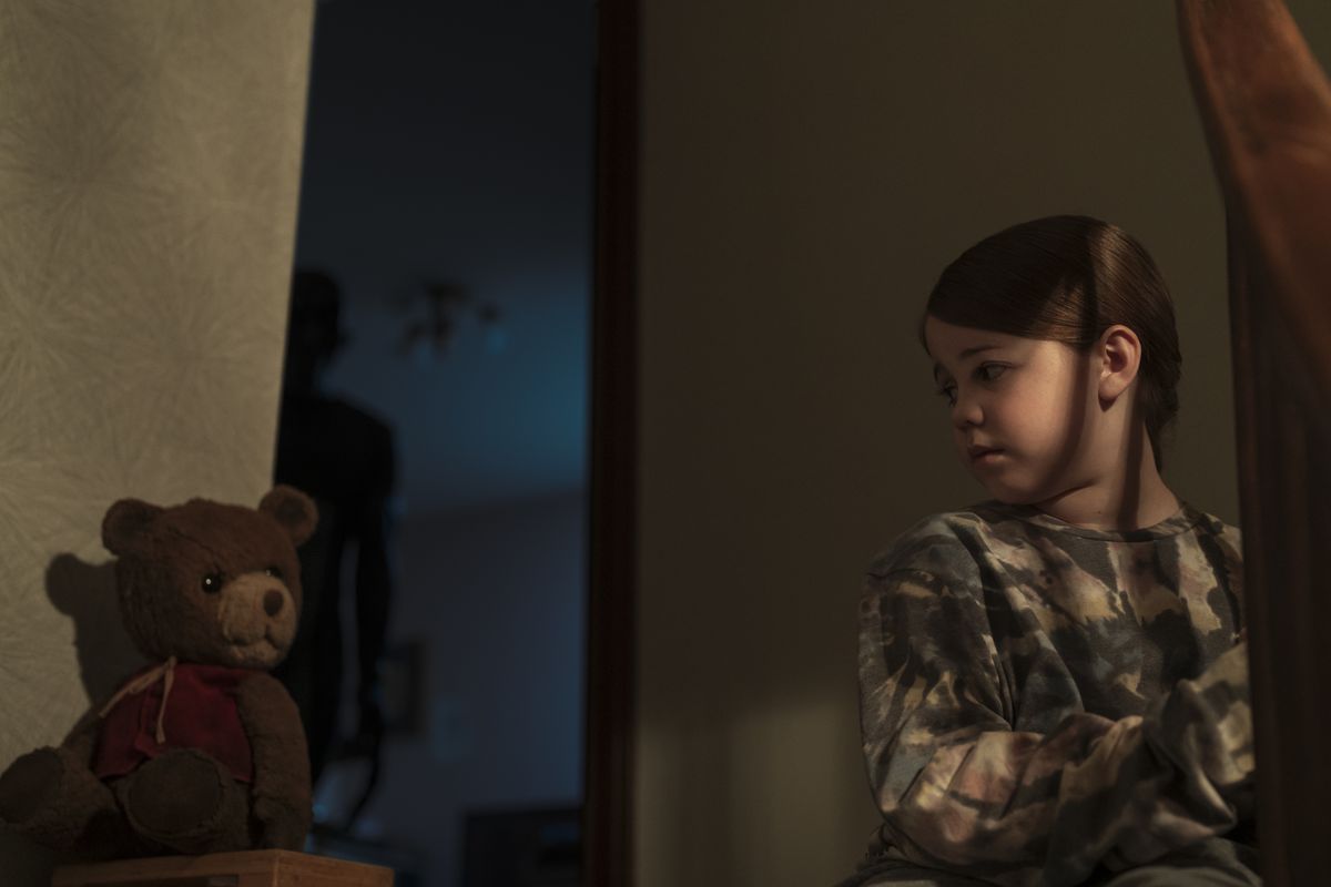 Une jeune fille jouée par Pyper Braun est assise en haut des escaliers à côté d'un ours en peluche tandis qu'une silhouette sombre et menaçante se cache derrière elle dans Imaginary.