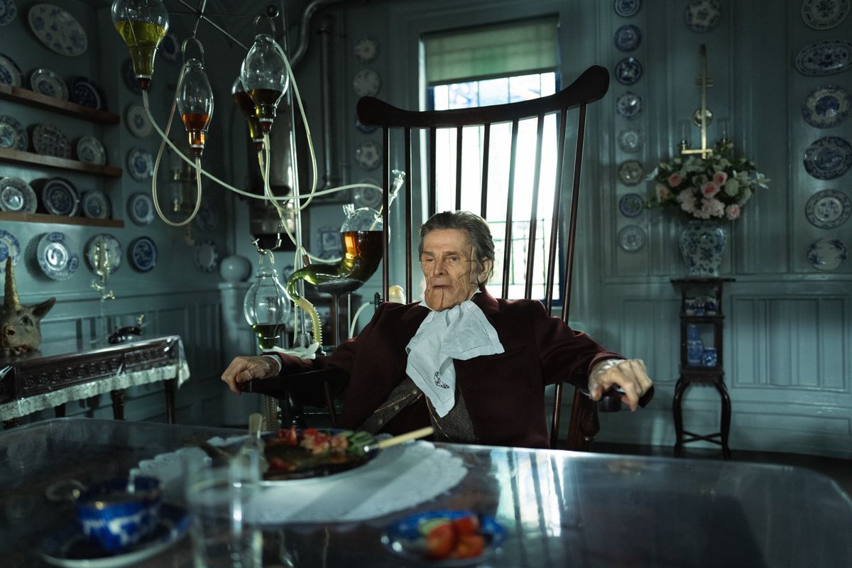 Willem Dafoe, au visage marqué, est assis sur une chaise à haut dossier avec à côté de lui un appareil en verre contenant divers liquides