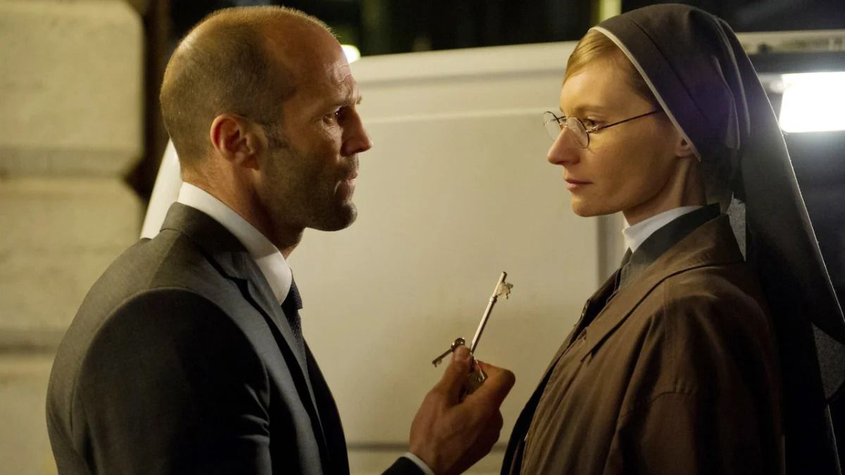 Un homme chauve en costume tient une paire de clés devant une femme avec des lunettes en habit de nonne.