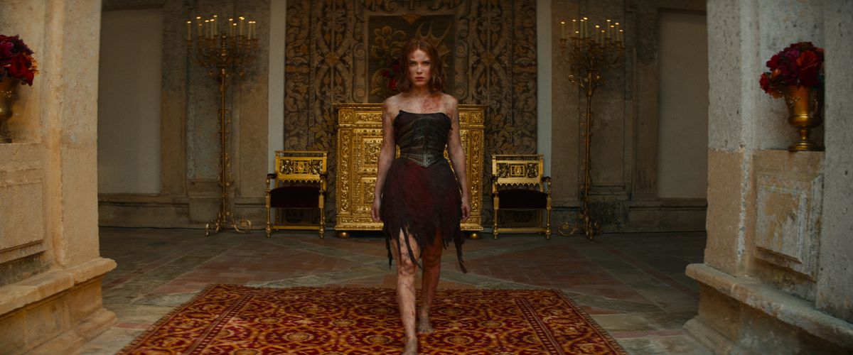 Elodie, jouée par Millie Bobby Brown, parcourt les couloirs dorés du palais dans une robe en ruine, déchirée et brûlée, la détermination sur le visage