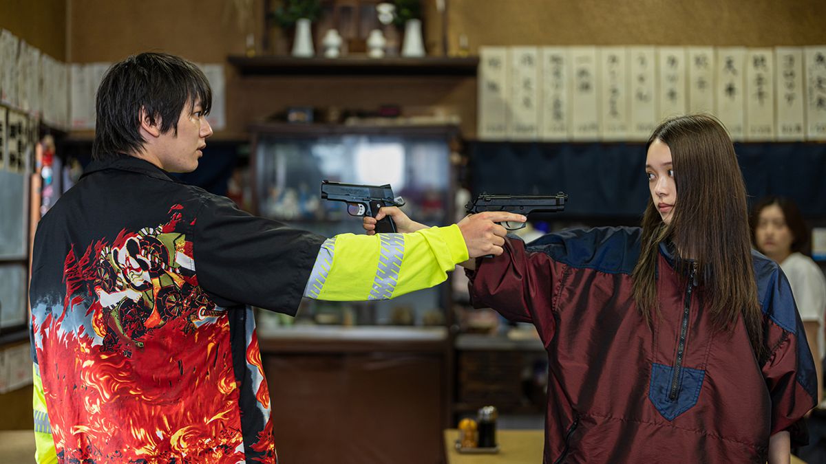 Akari Takaishi et un jeune homme vêtu d'une veste criarde se pointent des pistolets à l'intérieur dans Baby Assassins 2 Babies.