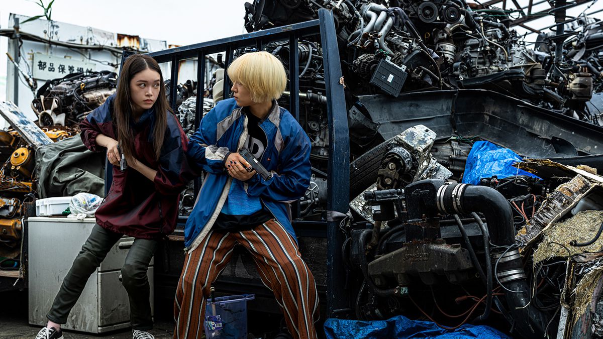 Akari Takaishi et Saori Izawa tiennent des pistolets et se cachent derrière une pile de pièces de voiture détruites dans Baby Assassins 2 Babies.