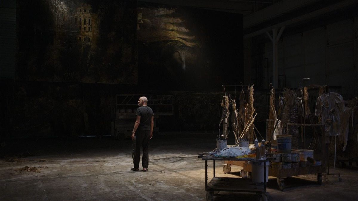 Un homme debout dans un studio sombre, entouré de fournitures artistiques et de grands tableaux.