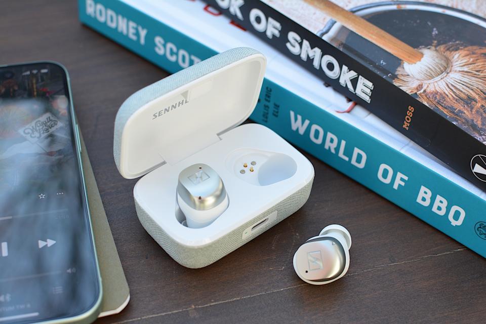 Écouteur Sennheiser Momentum True Wireless 4 blanc/argent posé dans son étui avec le couvercle ouvert, un écouteur posé sur une table en bois sur le côté.