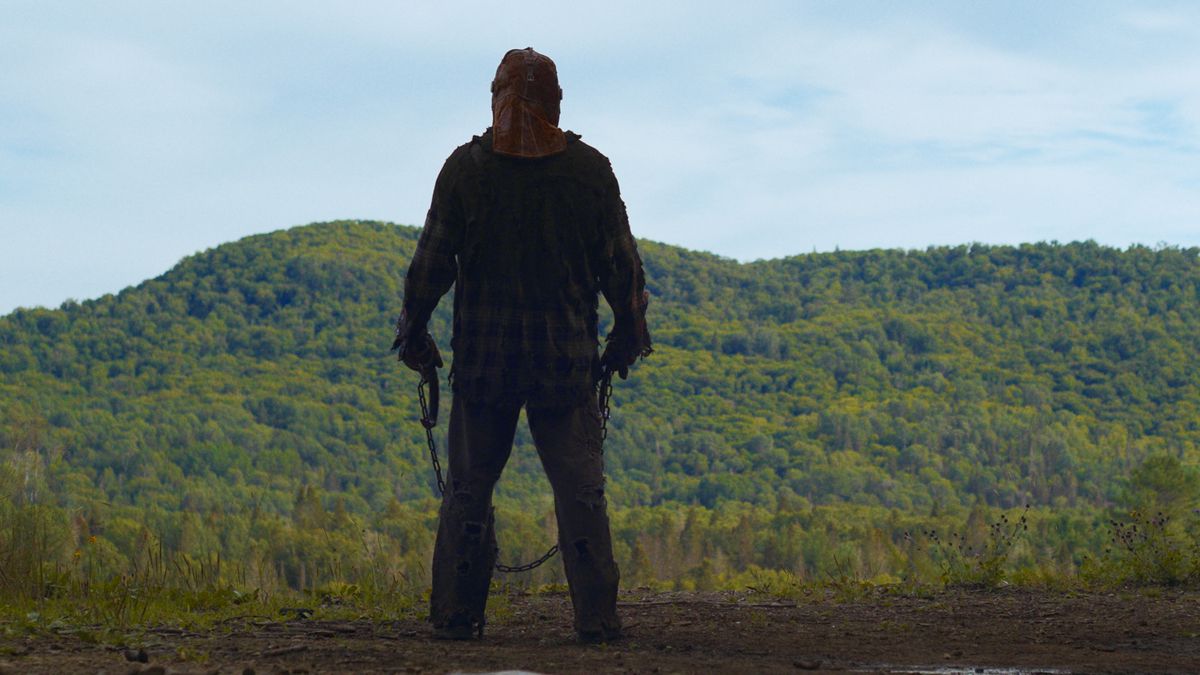 Un personnage avec un masque et deux crochets dans les mains se tient face à une forêt dans le film In A Violent Nature