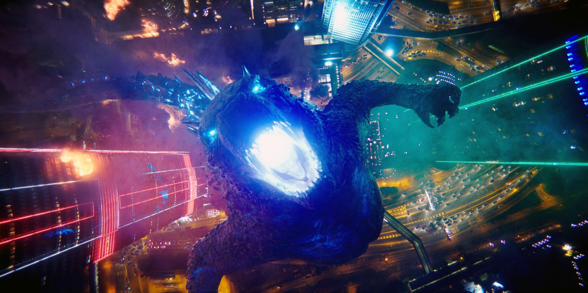 Une photo de haut en bas d'un Godzilla rugissant debout dans une ville éclairée au néon regarde droit vers son gosier bleu brillant dans Godzilla contre.  Kong