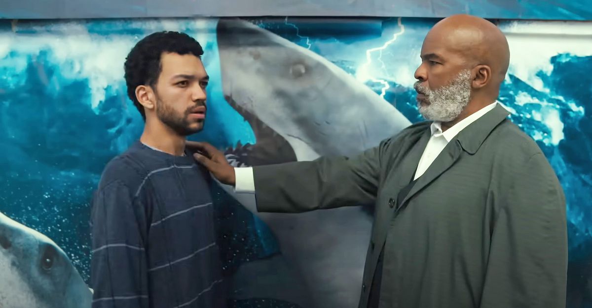 Un homme barbu plus âgé pose la main sur l’épaule d’un homme plus jeune devant une affiche de requin.