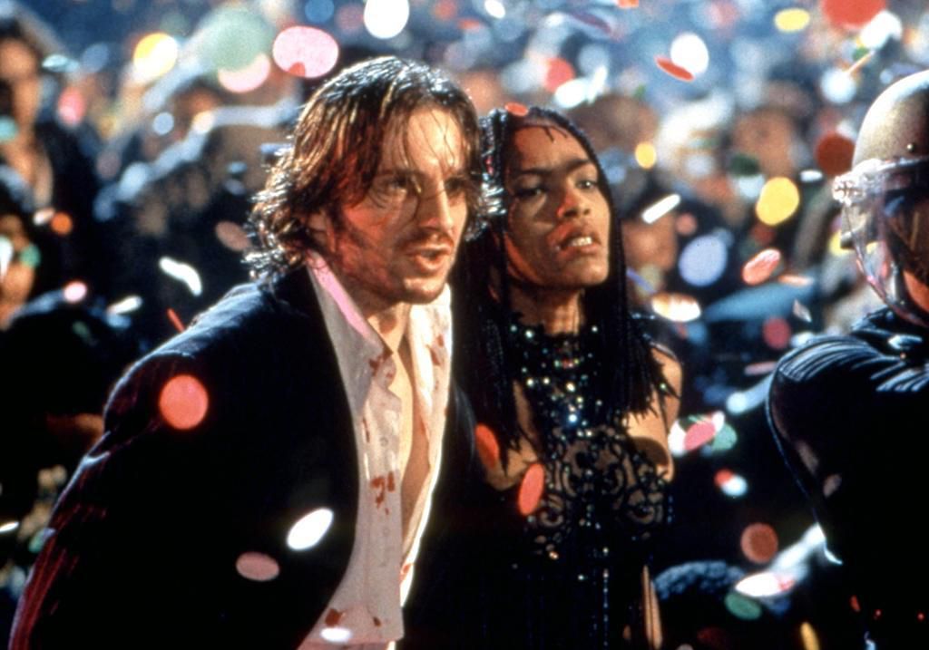 Un homme échevelé et une femme vêtue d’une robe noire se tiennent parmi une foule nombreuse tandis que des confettis pleuvent du ciel.