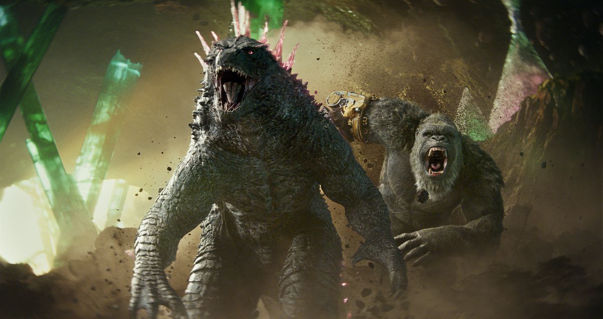 Godzilla, maintenant avec des pointes roses sur le dos, rugit tandis que King Kong, maintenant avec une main de robot, rugit derrière lui alors qu'ils se tiennent ensemble dans une caverne dans Godzilla x Kong : The New Empire.