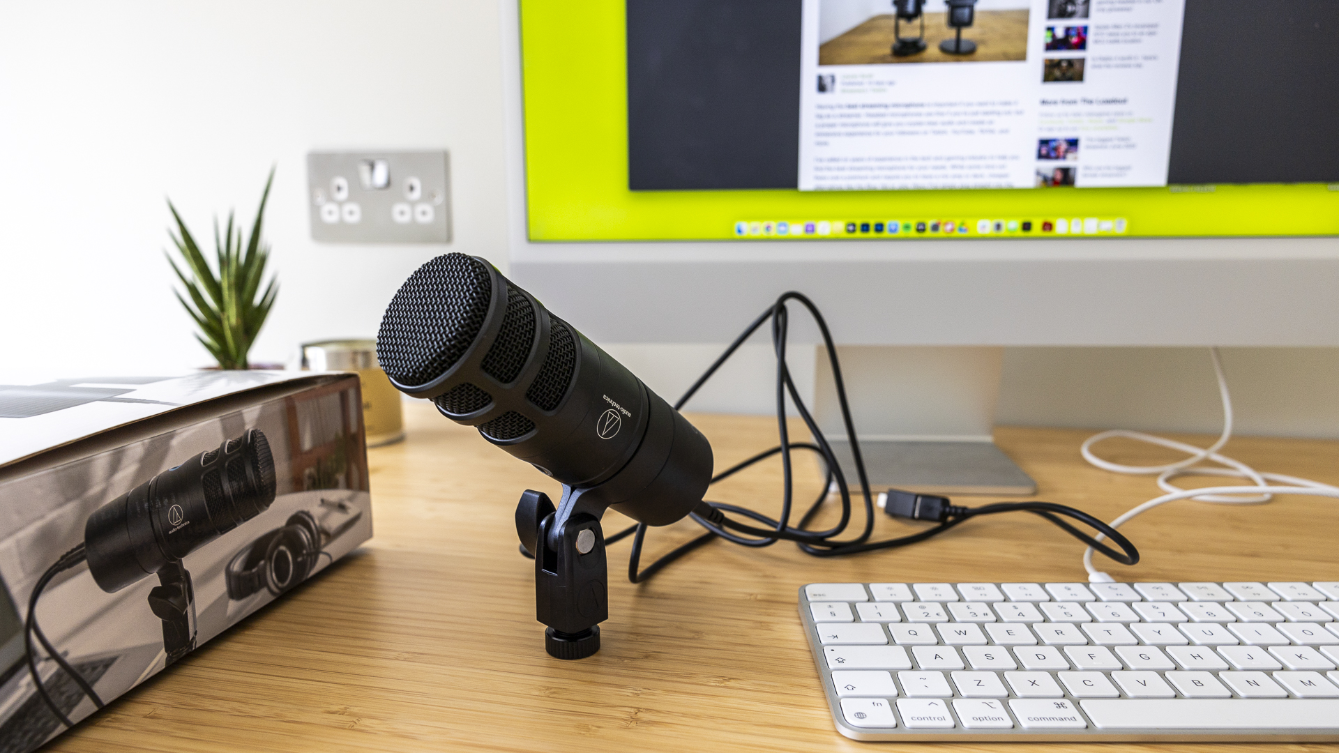 Image de revue Audio Technica montrant le microphone sur le bureau de quelqu'un à proximité d'une boîte et d'un clavier.